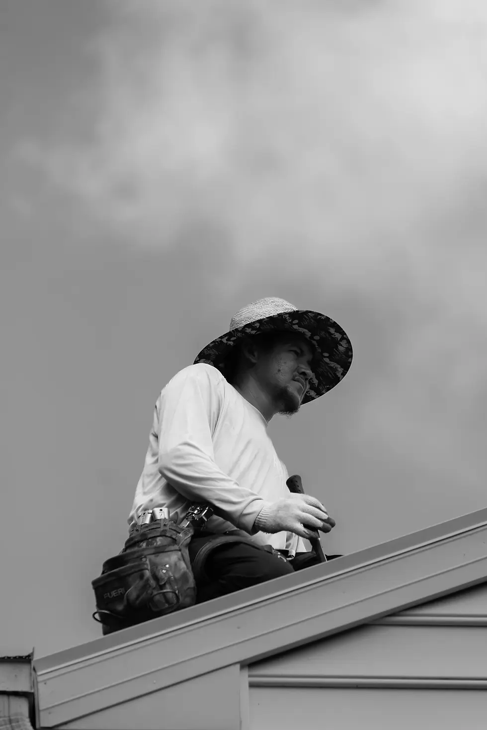 Black and white photo of roofer sitting on roof edge wearing wide brim hat tool belt and gloves holding tool with cloudy sky in background.