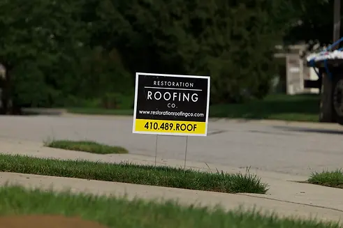 black-and-yellow-restoration-roofing-yard-sign-on-green-grass-near-sidewalk-promoting-comp