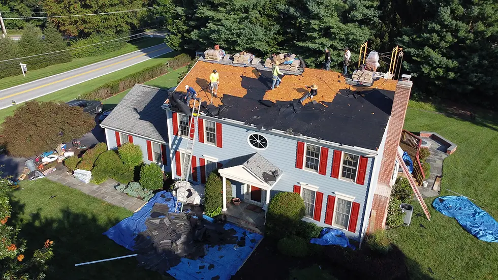Workers removing old shingles and underlayment on large two-story house with red shutters during construction phase showing roof replacement timeline with tarps covering yard and materials stacked on roof.