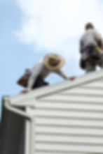 two-roofers-working-on-house-siding-one-wearing-straw-hat-and-gloves-the-other-standing-with-tool-belt-under-bright-sky