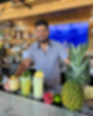 Employee smiling behind bar serving three tropical drinks with pineapple and fruit to the side