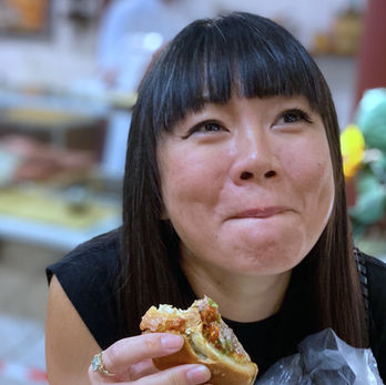 A woman with dark hair and bangs grins and looks upward as she holds her panino that is missing a few bites