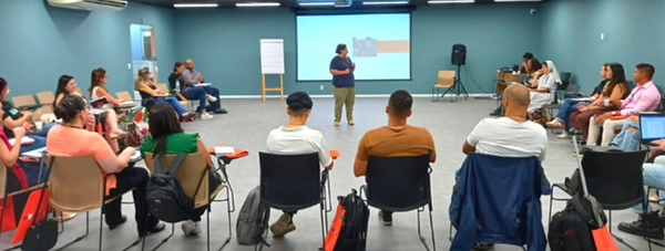 Carol Zanoti está em uma sala, ministrando o curso. Atras dela tem um projetor, a frente vários alunes sentados.