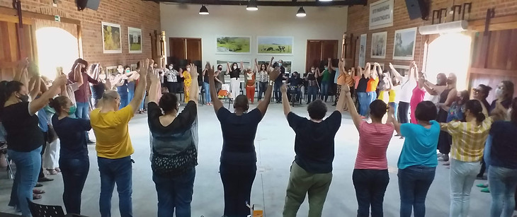 Dezenas de pessoas em roda em uma sala, estão de mãos dadas em direção ao alto