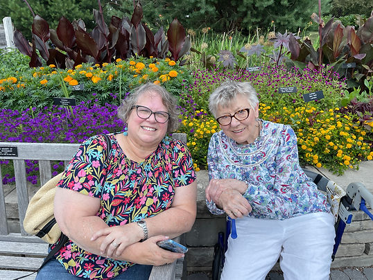 Two older women sittig in a flower garden leaning twoard one another and smiling at the camera