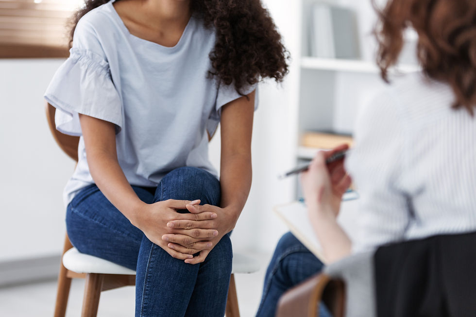 Two individuals seated; one holds a clipboard, in a counseling session. The setting is a bright, white room. One person's hands are clasped.