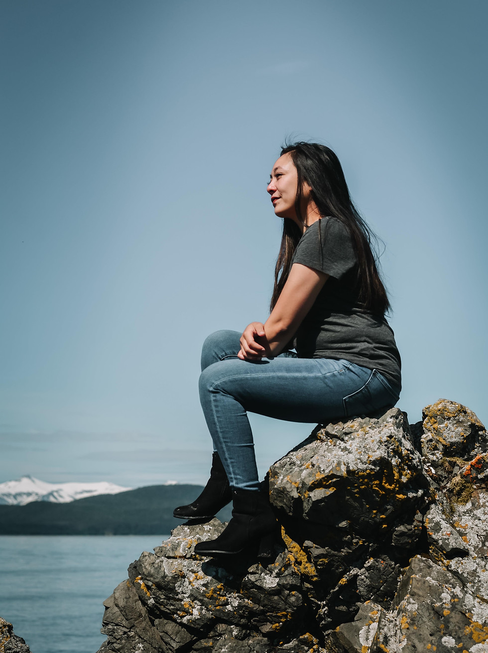 Rizza looking off camera and sitting on a rocky beach with water and mountains in the background.
