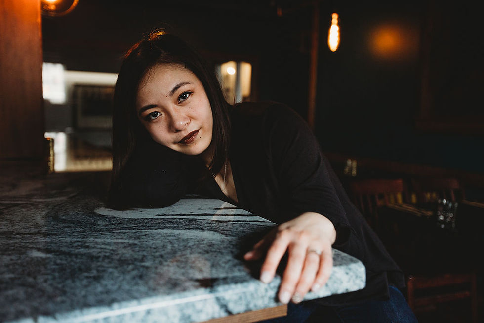 Rizza leaning on a table and looking at the camera in a darkly lit room.
