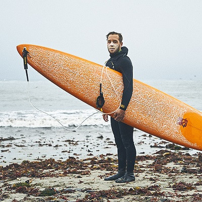 Full length shot of a man in black wetsuit standing on a seaweed-covered beach, holding an orange surfboard and looking directly at the camera.