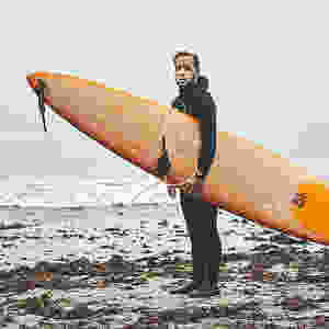 Full length shot of a man in black wetsuit standing on a seaweed-covered beach, holding an orange surfboard and looking directly at the camera.