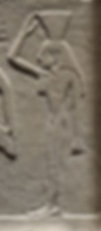 Ancient stone relief of a standing female figure raising an arm toward a triangular object resting on her head, on a weathered beige wall.