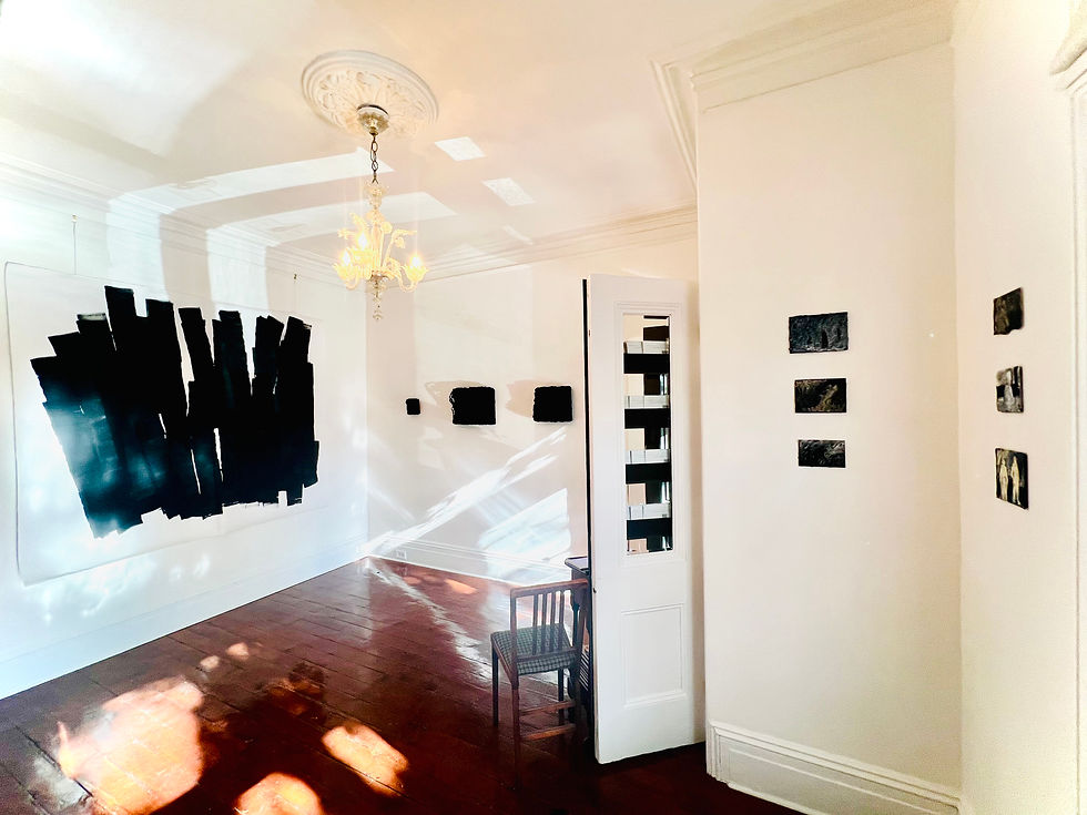 Art gallery room with abstract black paintings on white walls, a wooden chair, and a chandelier. Sunlight casts shadows on the floor.