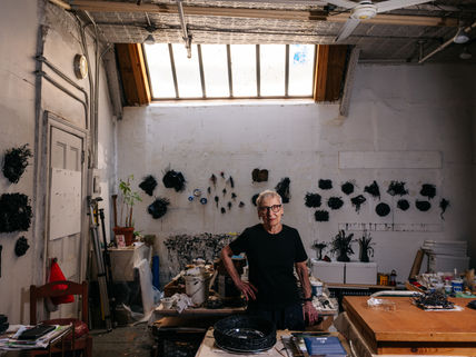 Gilda Pervin in her studio with black sculptures on walls, skylight overhead. Wooden tables, art supplies and plants around. Mood is creative and focused.
