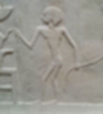 Ancient stone relief of a slender human figure in profile, holding a staff, carved on a beige wall with worn details.