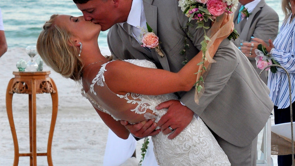 Bride and groom kiss passionately on a beach, she holds pink bouquet. He dips her in a gray suit, ocean and sunset in the background.