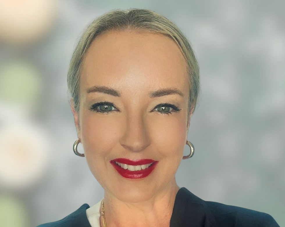 A person with short hair and red lipstick smiles warmly. They wear hoop earrings and a dark outfit. Blurred light background.