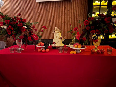 Opulent cake table styled with lush red florals, a tiered gold-accented cake, fresh fruit, and candles — set against a warm, moody backdrop for an intimate 50th birthday dinner.