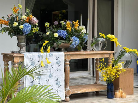 Rustic wooden console table styled with vibrant blue hydrangeas, yellow calla lilies, and assorted exotic florals, accented with taper candles, glass vases, and clustered candles — set against a garden backdrop for an elegant high tea celebration.