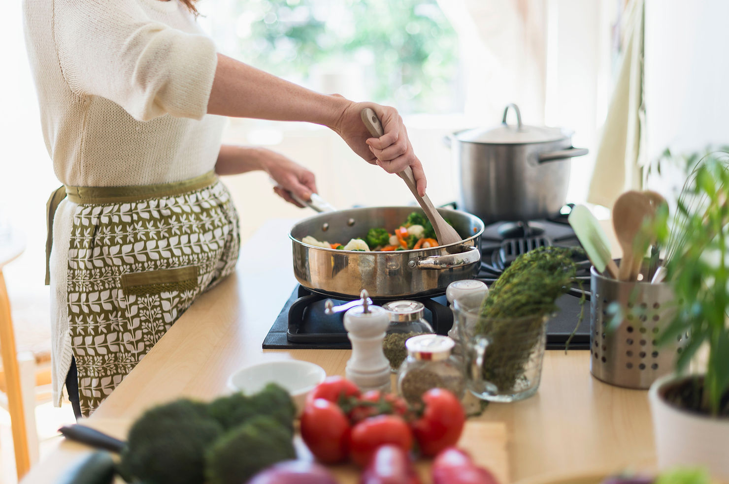 Person cooking vegetables in a pan on a stove. Kitchen setting with herbs, tomatoes, and utensils nearby. Bright, natural lighting.