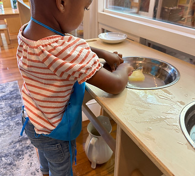 Toddler child in an orange and white striped shirt, wearing a blue apron, washes dishes at a toddler-sized sink at Oakland Community Montessori School, a preschool and Daycare in Oakland, California.