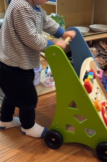 A toddler wearing a striped shirt and black pants pushing a green woodn walking support toy at Oakland Community Montessori School, a preschool and daycare serving children 18 months to 5 years old in Oakland and the East Bay