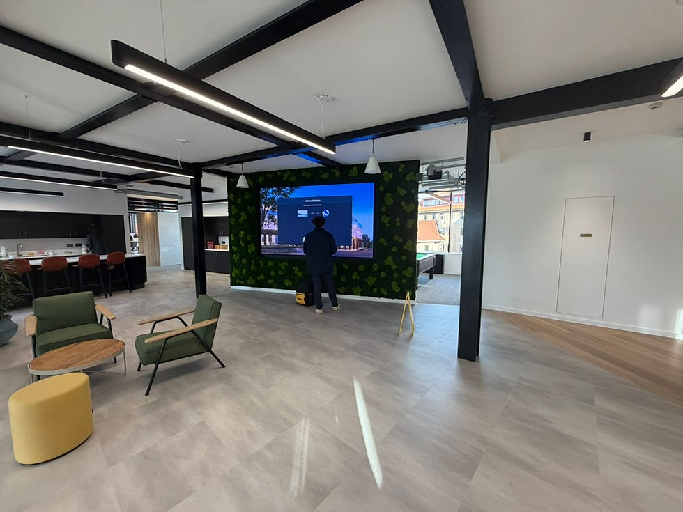 A person stands at a large screen on a leafy wall in a modern open office with green chairs and wooden accents. Another person is by the counter.
