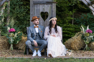Mariés alsaciens posant assis devant une cabane entourée de fleurs pendant une cérémonie laïque.