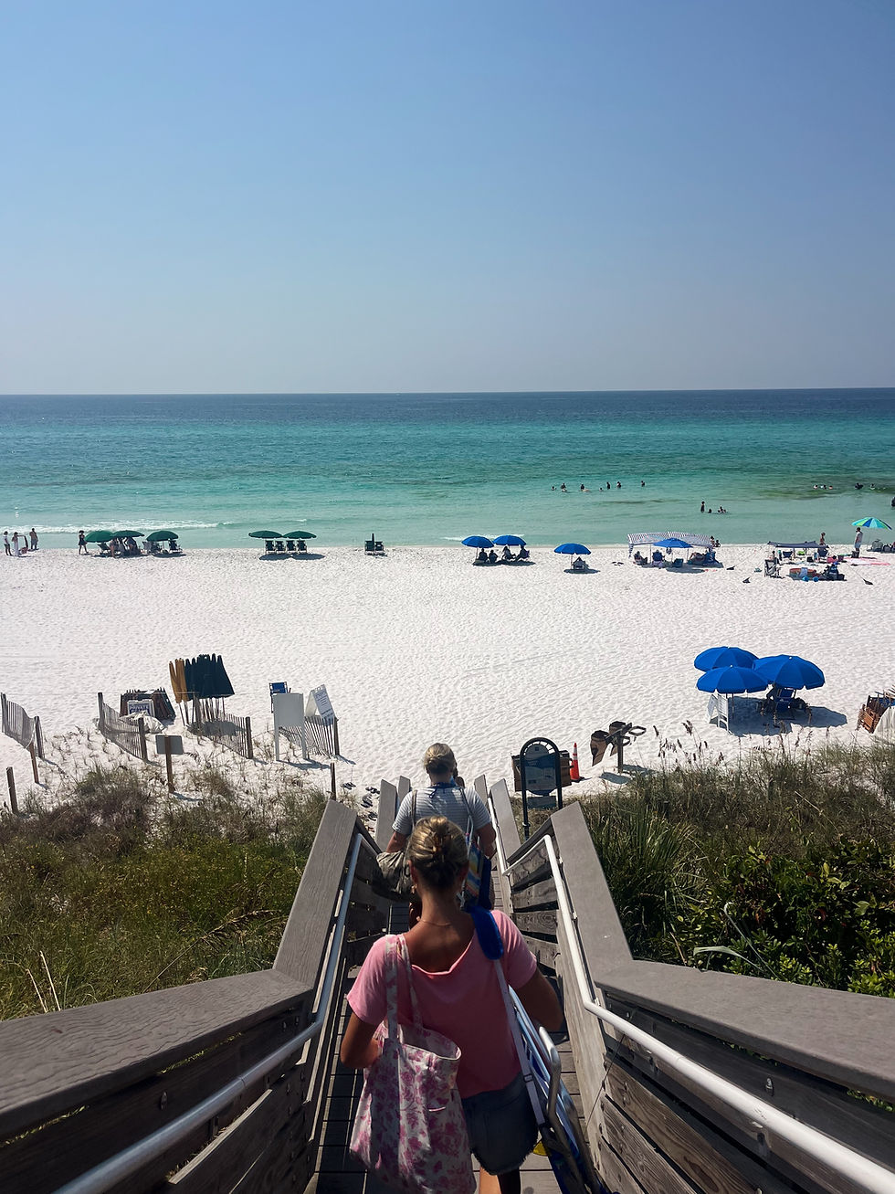 Broad view of a beach with blue umbrellas and chairs scattered around and a blue ocean washing up on the shore. Few people standing around in the water.