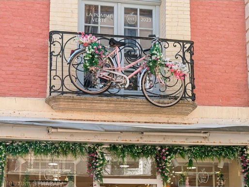 A brightened photo of a European storefront with a pink brick façade and cream-colored stone pillars. On a black wrought-iron balcony on the second floor, a pink vintage bicycle decorated with pink and white flowers is mounted.
