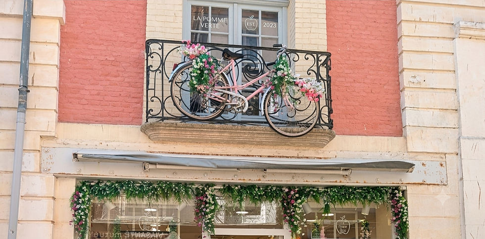 A brightened photo of a European storefront with a pink brick façade and cream-colored stone pillars. On a black wrought-iron balcony on the second floor, a pink vintage bicycle decorated with pink and white flowers is mounted.