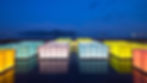 Illuminated rectangular pavilions in various colors including yellow, pink, blue, and green floating on a shallow water basin at dusk, with mountains and ships visible across the water in the background
