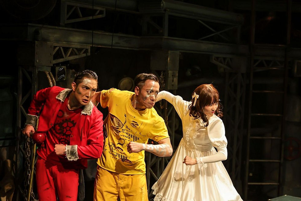 GEAR Kyoto performer in white dress as the Doll character standing between two Roboroid performers in red and yellow costumes on a dark industrial stage