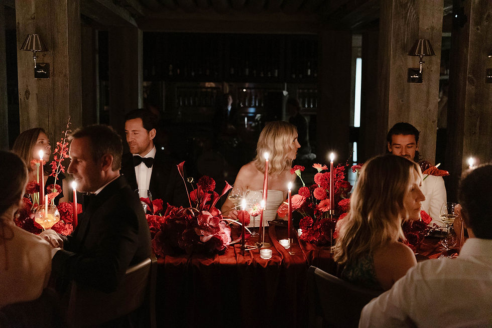 Candlelit formal dinner with guests in tuxedos and dresses around a table of red flowers in a dark wood room.
