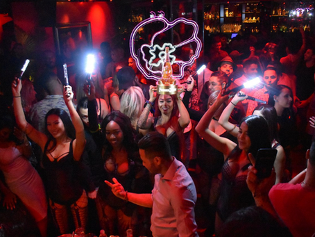 People dancing in a dimly lit club, holding lights and a champagne bottle with neon lights overhead. The mood is lively and festive.