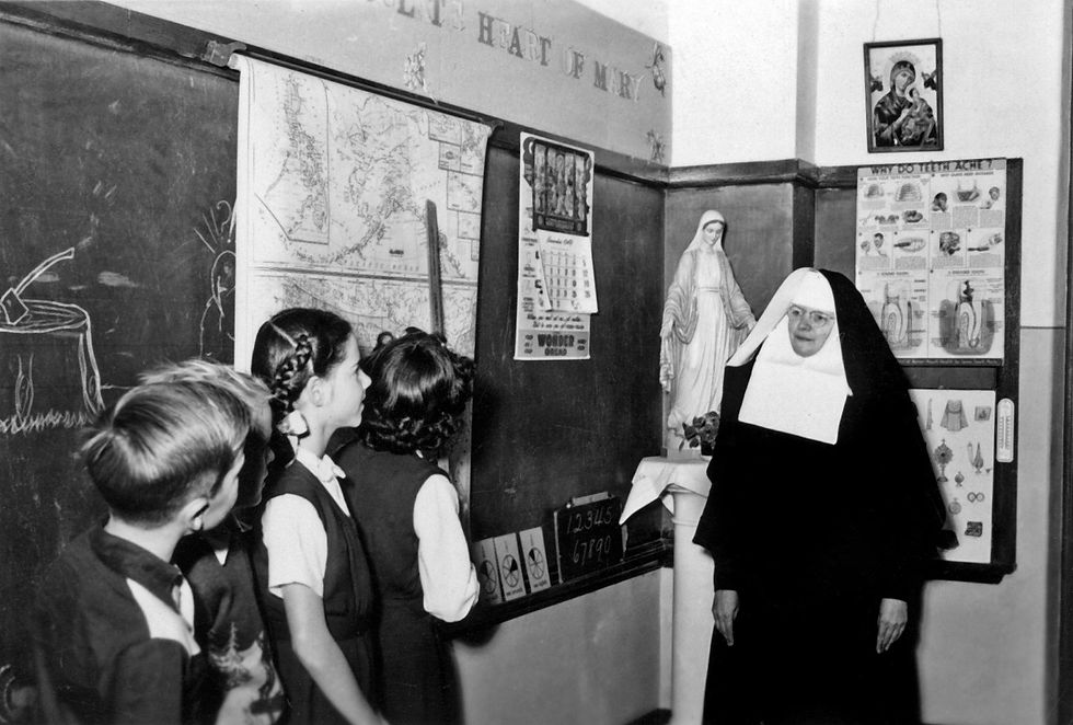 A Catholic sister in full habit stands beside a chalkboard and a statue of Mary, teaching a group of young students in a mid-20th century classroom.