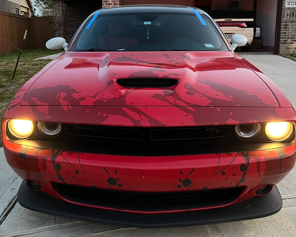 Front view of a red car with custom hood graphics, headlights on, in a driveway at dusk. Landscape orientation, car centered, dominant color red.