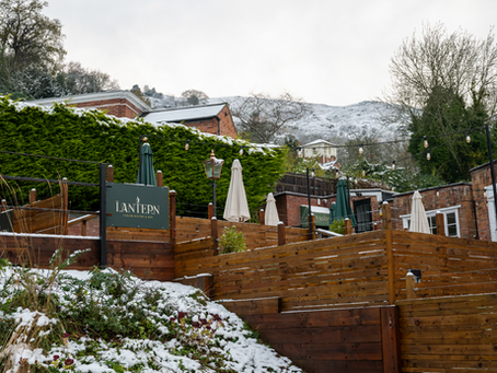 Snow-covered outdoor terrace of The Lantern Pub & Kitchen in Malvern Hills with wooden fencing and green umbrellas.