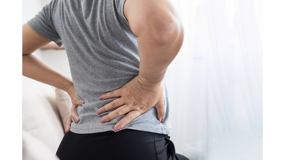 Person in a gray shirt holds lower back in pain and discomfort. Soft background with sheer curtains. 