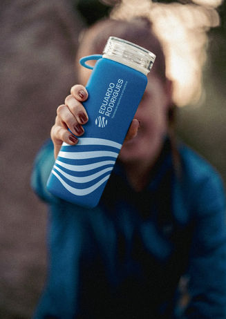 Uma pessoa segura uma garrafa de água esportiva de cor azul fosco. A garrafa é personalizada com o logotipo branco no topo e o padrão de ondas fluidas na base, reforçando o branding para o público atleta.