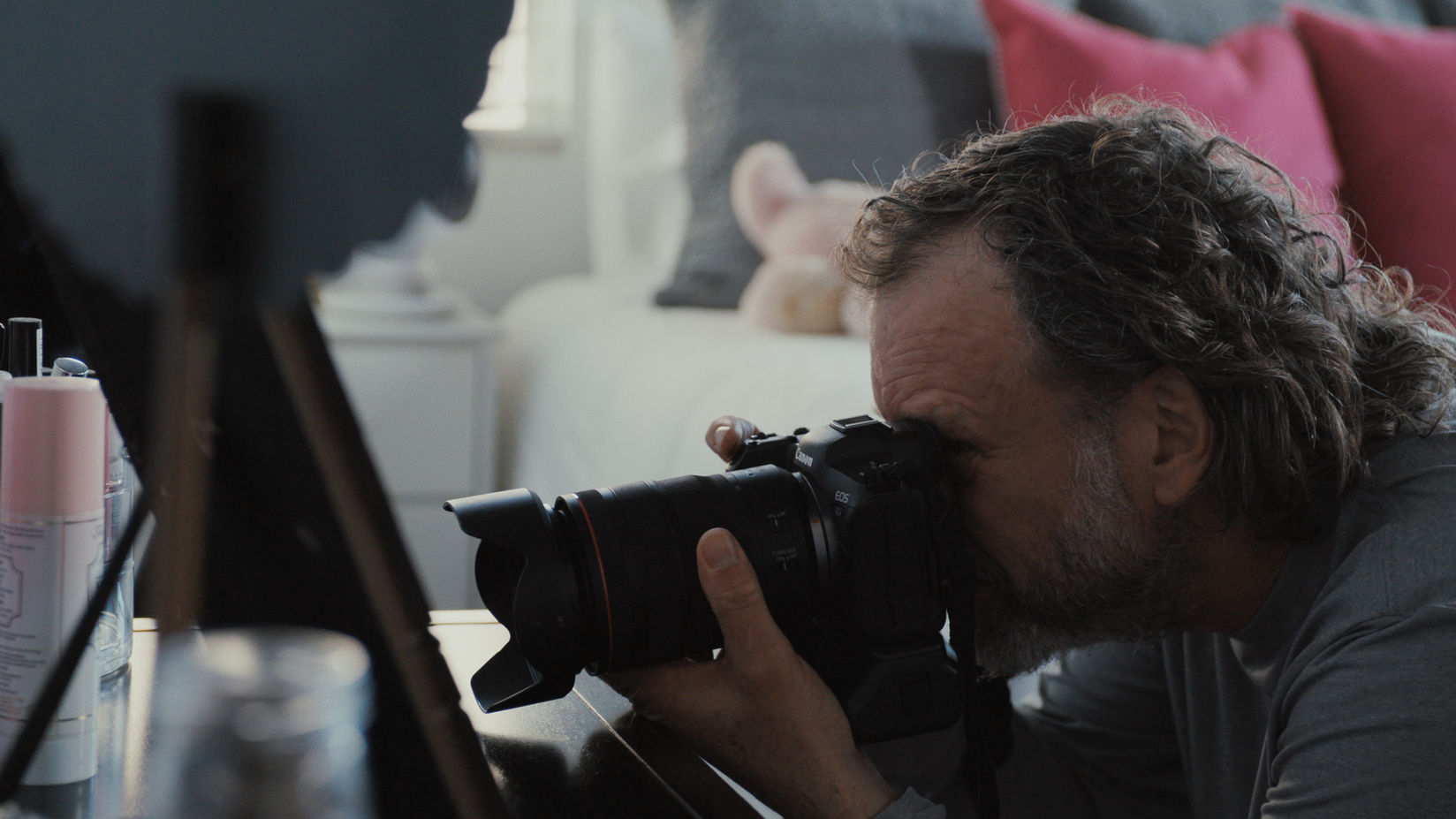 Photographer Lou Bopp crouches with his camera aimed at a surface in a brightly lit bedroom.