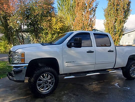 Lifted white Chevy Silverado 2500HD pickup on a wet driveway with fall trees behind it