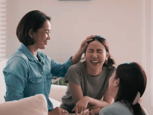 An Asian mother sitting on a couching laughing with her two teen daughters.