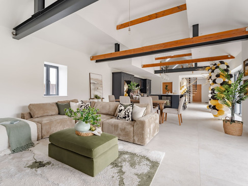 Open-plan living area with exposed beams, large sectional sofa, green ottoman, dining space and decorative balloon arch near patio doors.