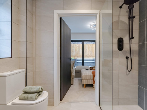 Modern bathroom with a glass-enclosed shower and a neat stack of green towels on a toilet. Through an open door, a cozy living area is visible, highlighting a serene ambiance.