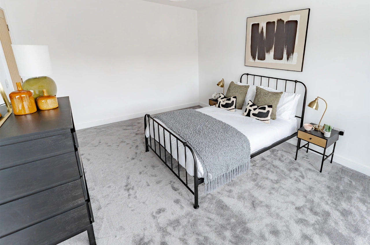 Minimalist bedroom with a double bed featuring a gray throw and patterned pillows, flanked by gold lamps. Dark dresser with decorative vases, abstract wall art.