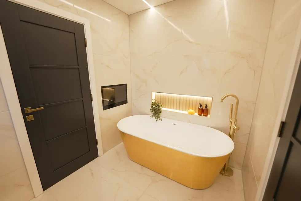 Luxurious bathroom with a gold bathtub, beige marble walls, and black door. Backlit shelf with plants and bottles creates an elegant vibe.