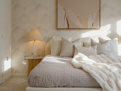 Cozy bedroom with a large, neatly-made bed covered in plush pillows and a soft blanket. Warm lighting from bedside lamps creates a serene, inviting atmosphere.