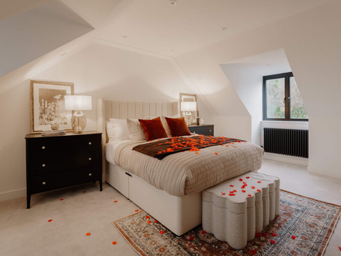 Cozy bedroom with a plush bed, red and cream cushions, and rose petals. Soft lighting from table lamps, elegant black side tables, and an ornate rug.