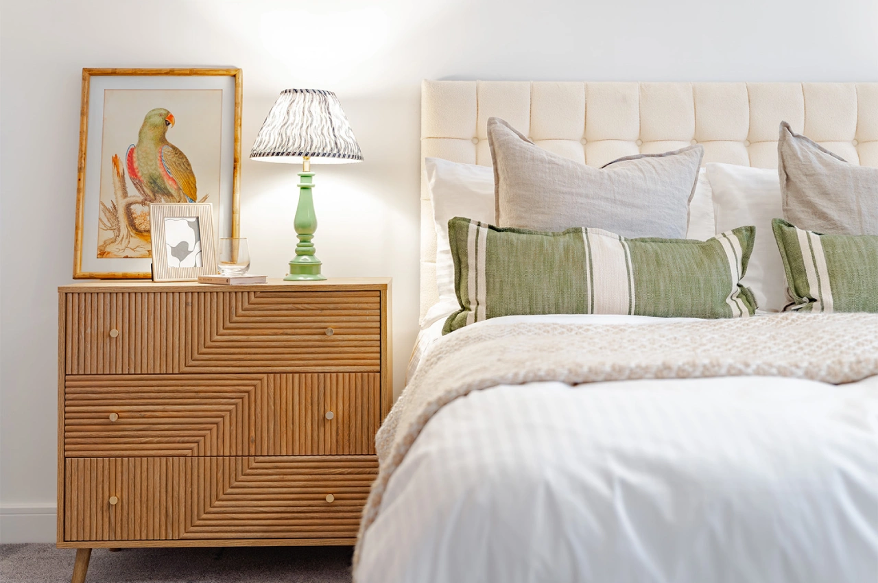 Cozy bedroom scene featuring a cream upholstered bed with green pillows, a wooden nightstand with chevron patterns, framed parrot art, and a lit lamp.