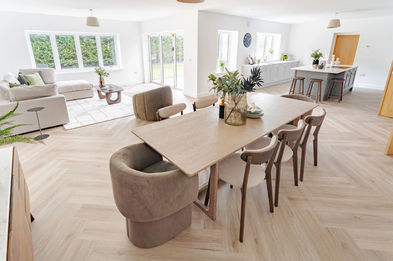 Spacious, modern open-plan living area with neutral tones. Features a wooden dining table, cozy sofa, large windows, and a kitchen island. Calm atmosphere.